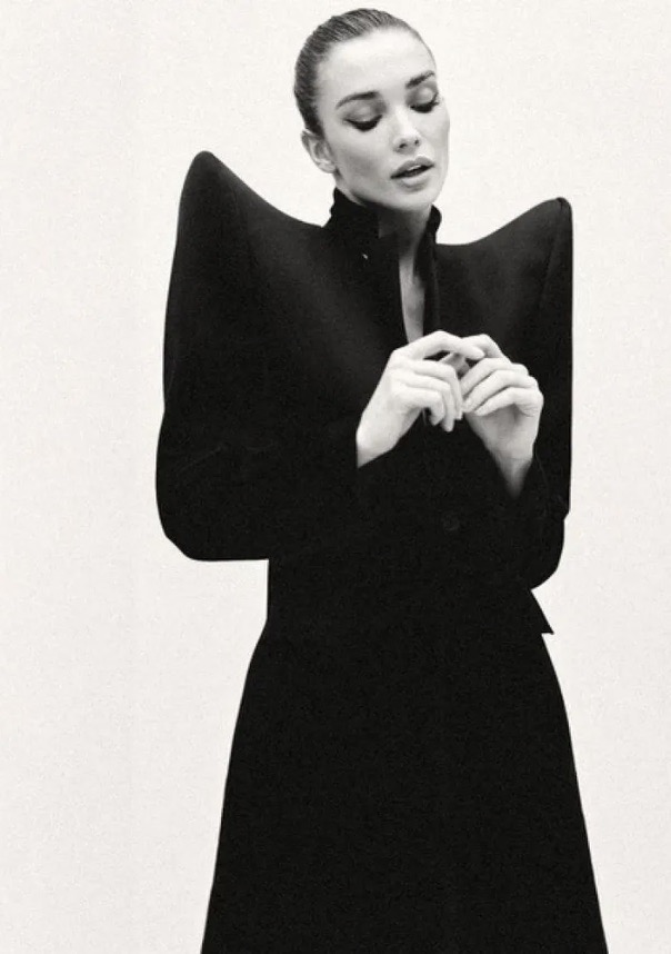 Black and white portrait of a woman in a black coat with exaggerated shoulder pads against a white background in a studio setting.