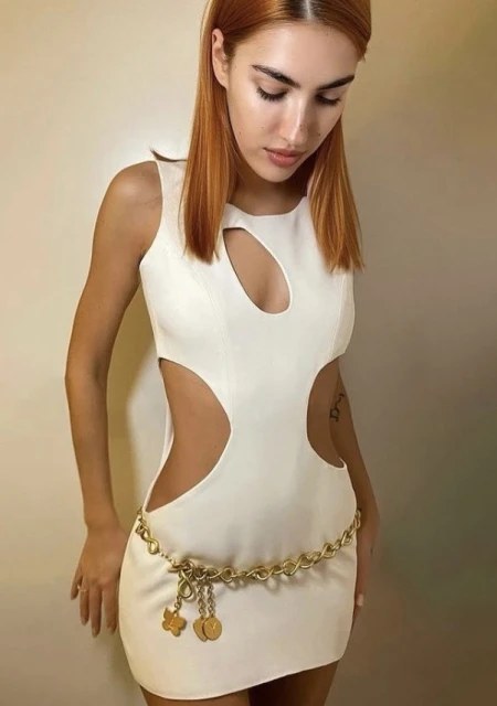 A woman with auburn hair poses indoors wearing a white cutout dress and a gold chain belt with dangling charms.