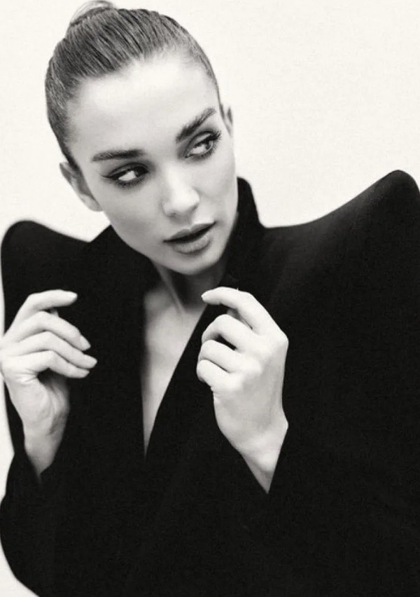 Monochrome portrait of a woman with dark hair in a bun wearing a structured black jacket against a plain background