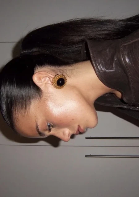 Profile of adult female with slicked-back hair wearing dark brown leather jacket and gold earrings with black gemstones in an indoor setting.