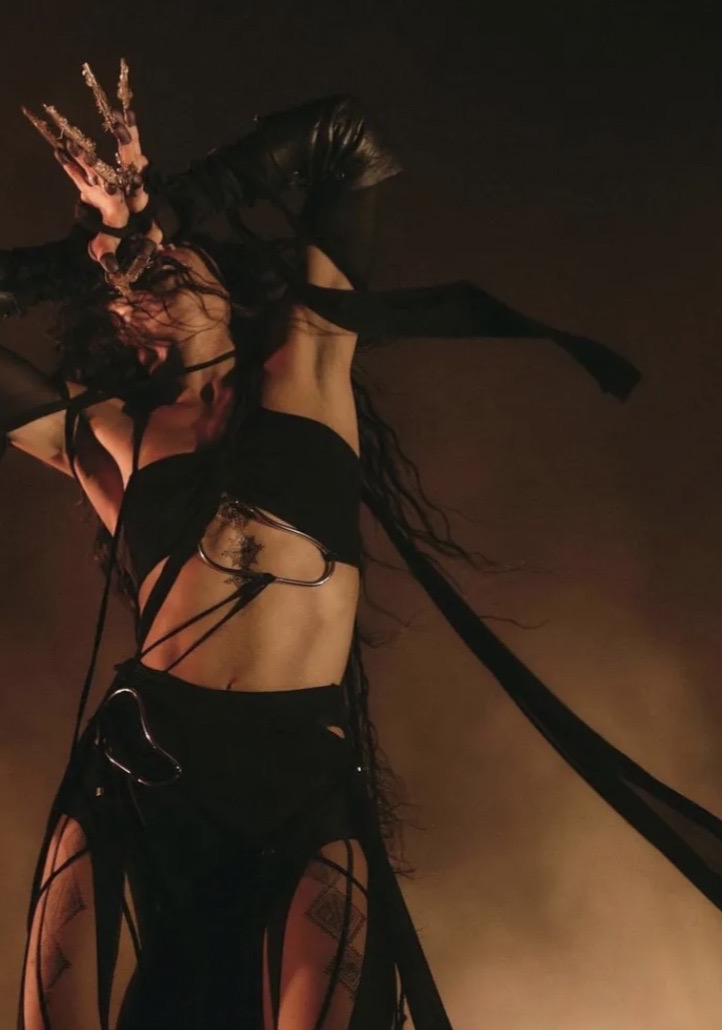 Woman in a dark setting wearing a black costume with a detailed headdress, jewelry, and flowing fabric accentuating a dramatic pose.
