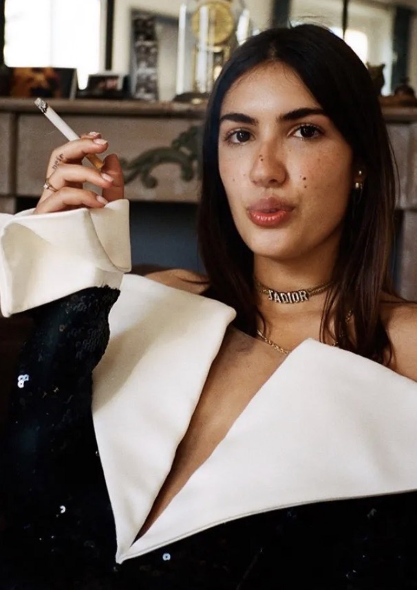 Brunette woman with fair skin poses indoors holding a cigarette, wearing an off-shoulder black and white dress and a gold necklace.