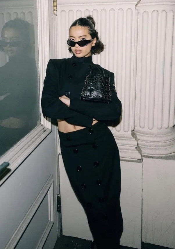 A fashionable woman poses indoors in a stylish black two-piece outfit and sunglasses, holding a matching black purse, for a fashion shot.