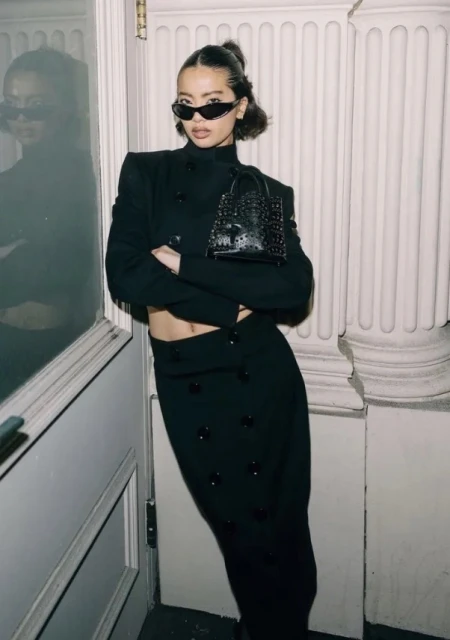 A fashionable woman poses indoors in a stylish black two-piece outfit and sunglasses, holding a matching black purse, for a fashion shot.