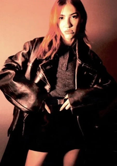 A young adult female with orange hair poses in a photo studio, wearing a black leather jacket, grey shirt, and black skirt. 