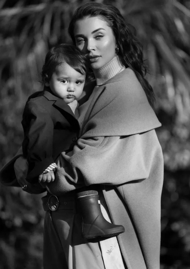Monochrome portrait of a woman holding a child in a coat outdoors, showcasing a fashion-editorial style with shallow depth of field.