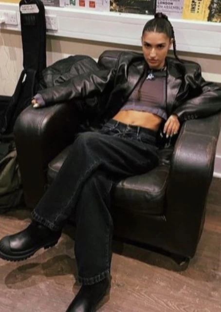 A woman is seated in a black armchair wearing a leather jacket, crop top, and black jeans in an indoor setting.