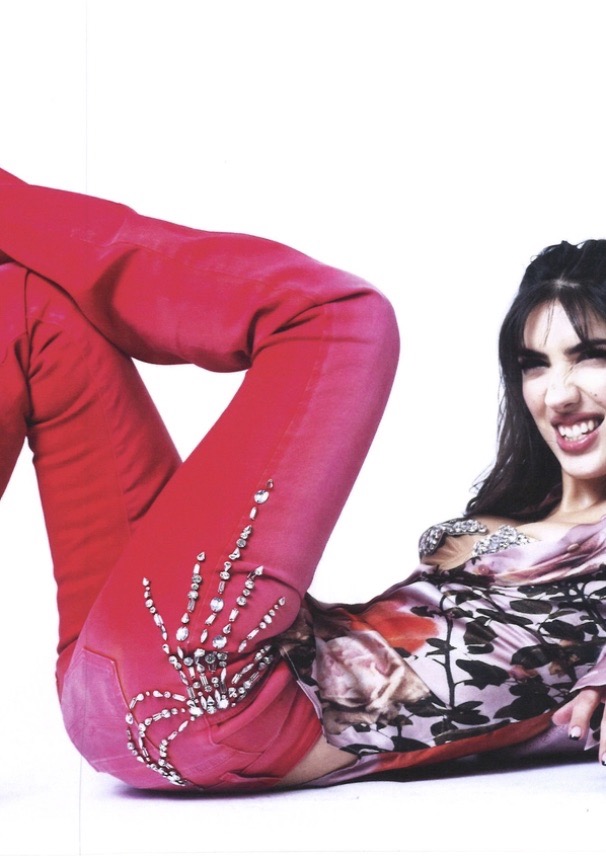 An adult female laying with knees up on white backdrop wearing an embellished floral top and embellished pink pants in a studio shot.
