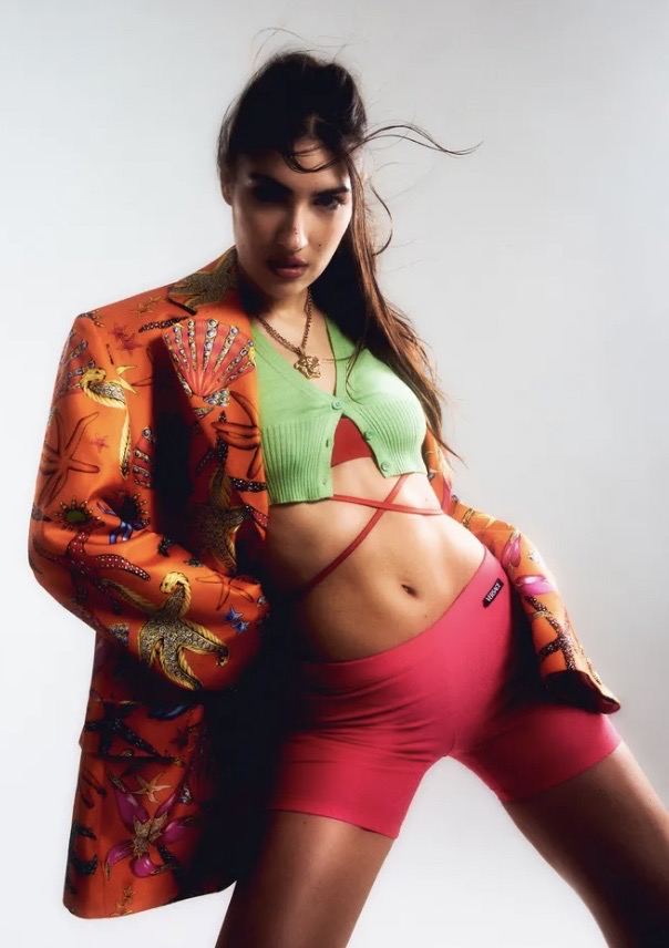 Portrait of a young woman in a patterned blazer, knit top, and athletic shorts against a white background in a fashion editorial.