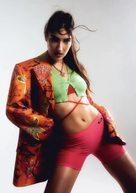 Portrait of a young woman in a patterned blazer, knit top, and athletic shorts against a white background in a fashion editorial.