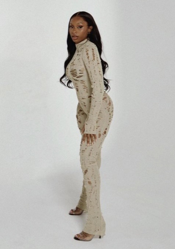 A slender woman with long brown hair poses in a ripped beige jumpsuit and high heels against a white background in a fashion style shot.