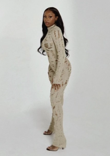 A slender woman with long brown hair poses in a ripped beige jumpsuit and high heels against a white background in a fashion style shot.