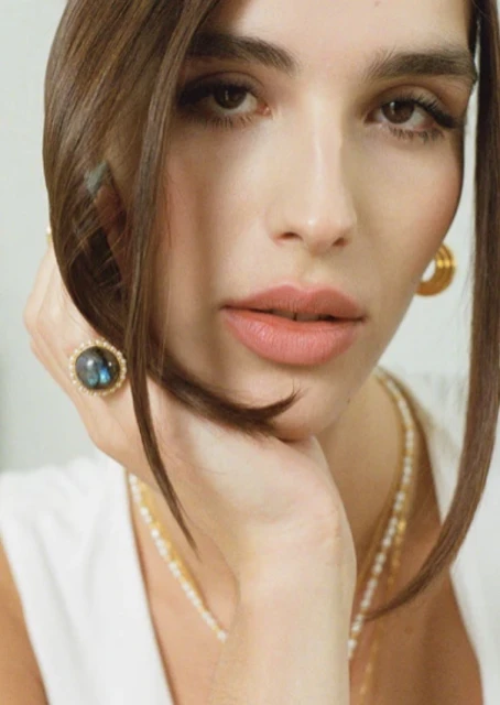 Close-up portrait of a woman with dark hair wearing pearl and gold jewelry and a large, dark ring with an ornate gold setting.