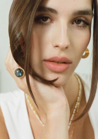 Close-up portrait of a woman with dark hair wearing pearl and gold jewelry and a large, dark ring with an ornate gold setting.