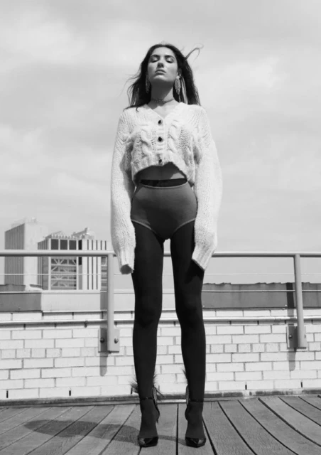 Black and white shot of a woman in a cropped knit sweater, shorts, and boots standing on a wooden deck against a sky background.