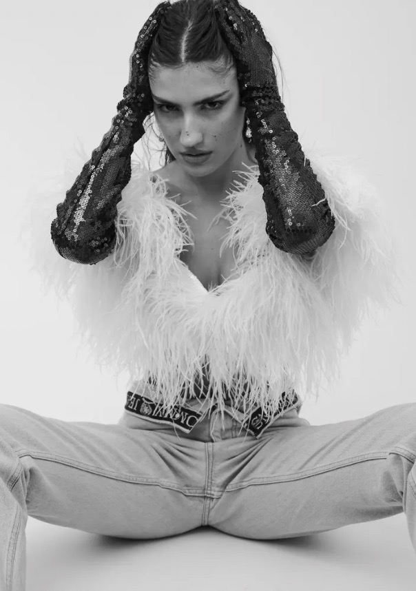 Monochrome shot of a woman in jeans, feather jacket, and sequin gloves striking a pose against a white backdrop in a studio setting.