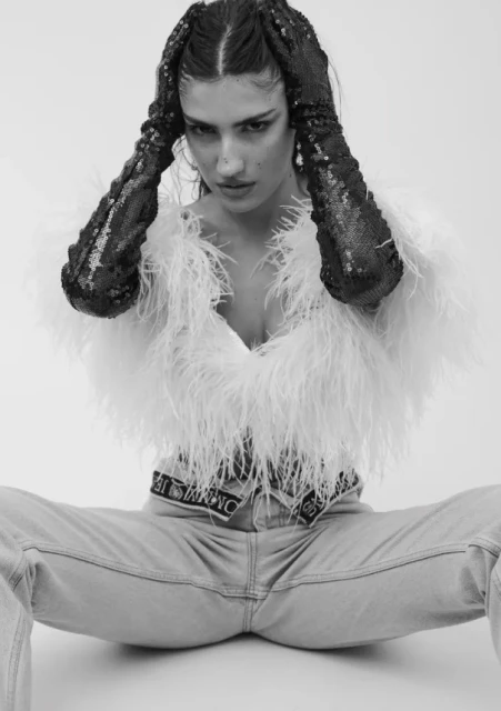 Monochrome shot of a woman in jeans, feather jacket, and sequin gloves striking a pose against a white backdrop in a studio setting.