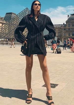 Model wearing a black pinstripe blazer and dark sunglasses standing in a public square with a glass pyramid background.