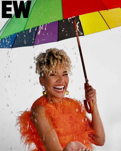 A smiling woman in an orange feathered dress stands under a rainbow umbrella in the rain, looking at the camera, with a white background.