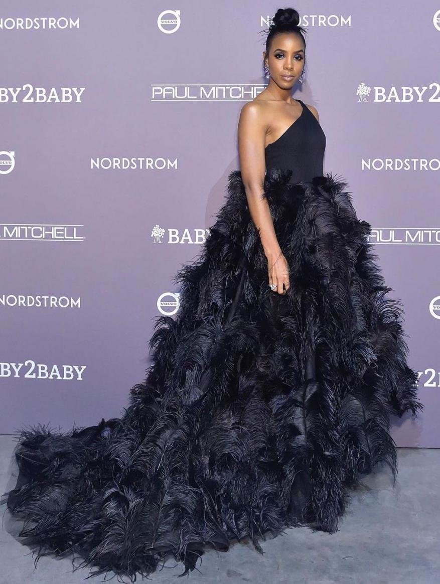 Kelly Rowland poses on a red carpet in a one-shoulder black dress with a large feathered skirt, attending the Baby2Baby charity gala.