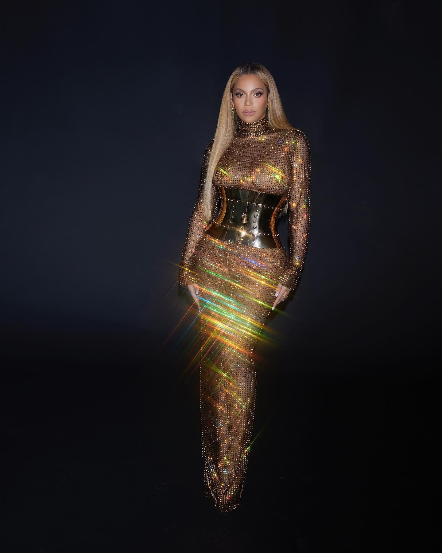 Beyoncé poses in a shimmering gold, floor-length gown with a wide gold belt against a dark background, radiating elegance and style.