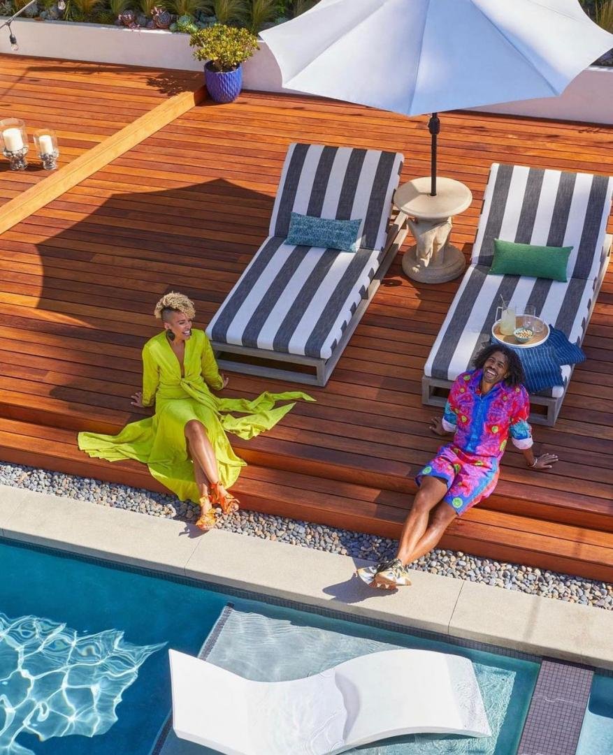 Two women relax by the pool on a sunny day on a wooden deck with lounge chairs and a white sun umbrella overhead creating shade.