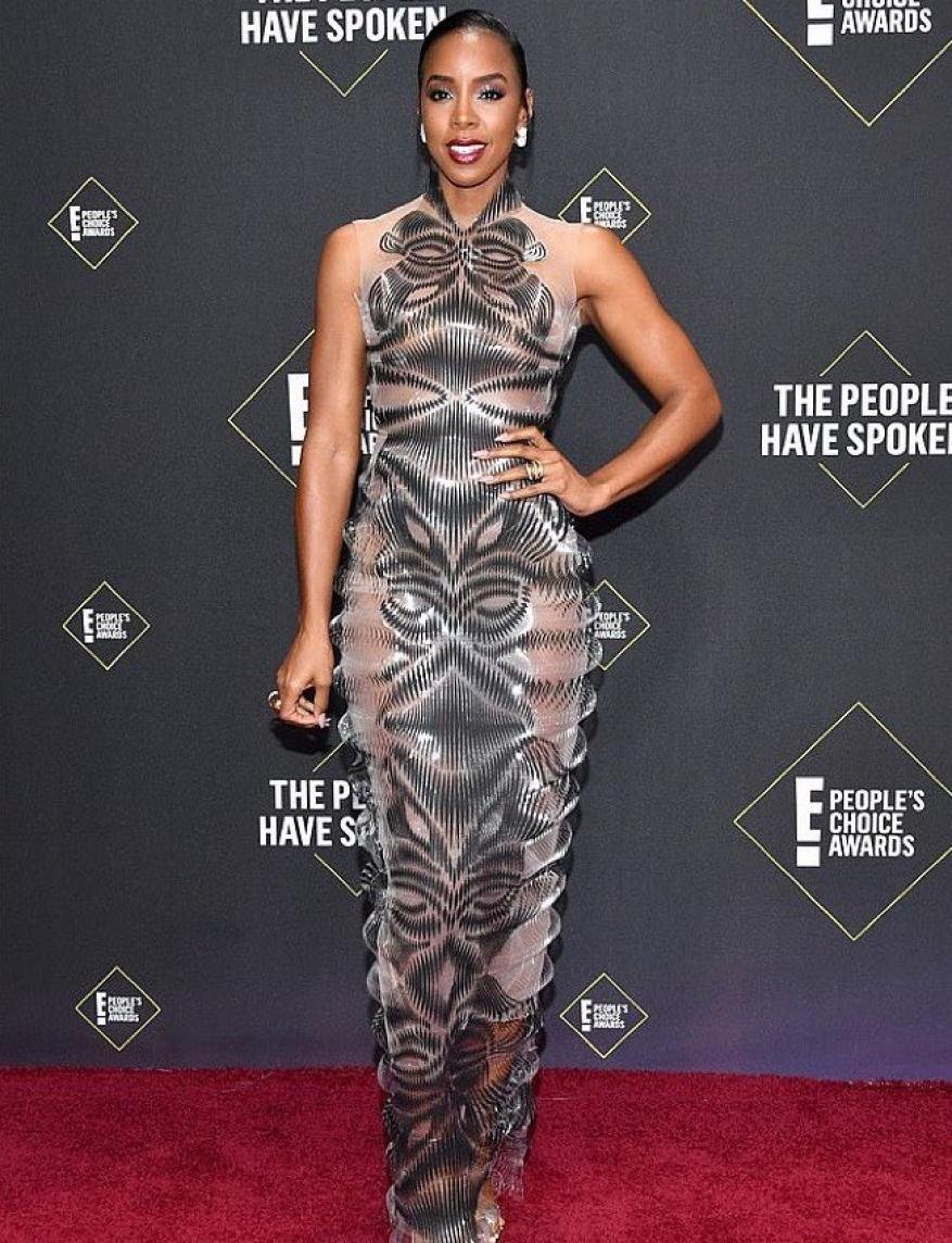Kelly Rowland poses on a red carpet in a form-fitting, grey and silver gown with a high neck and arm on hip, smiling at the camera.