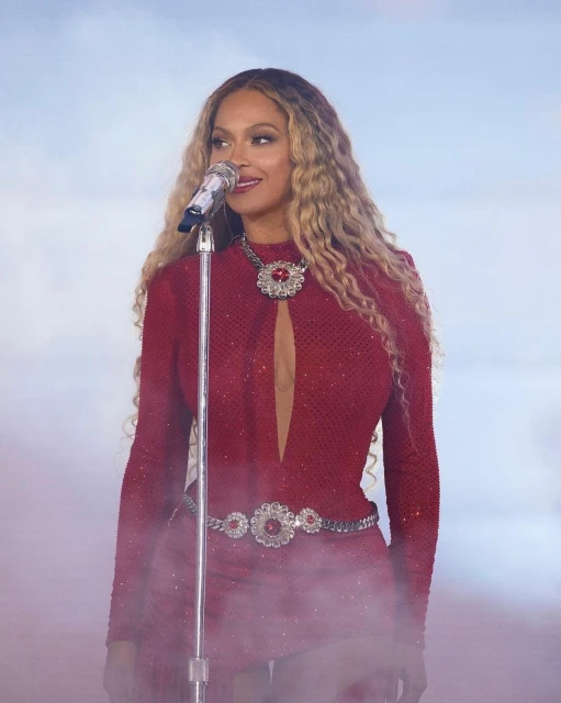 Beyoncé performs on stage wearing a red sequined dress with embellished necklace and belt, singing into a microphone with curly hair.