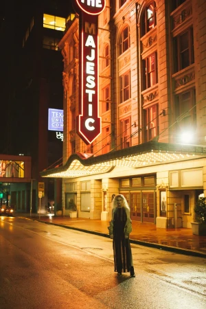 Night shot of a person standing on a wet street in front of The Majestic theater with its neon sign illuminated at night.