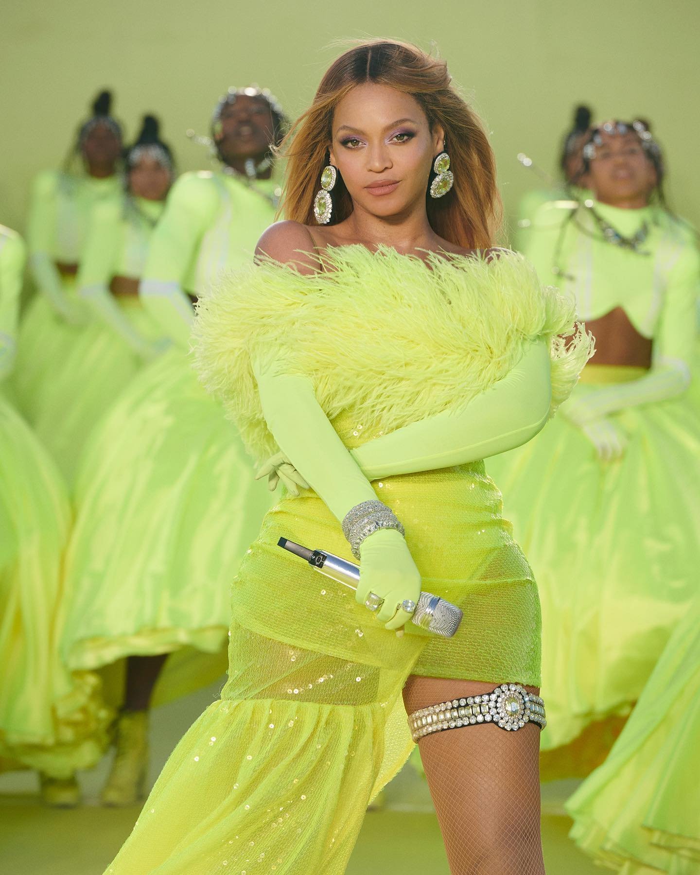 Singer Beyoncé performs in a yellow feathered dress, holding a microphone, with dancers in matching outfits in the background.