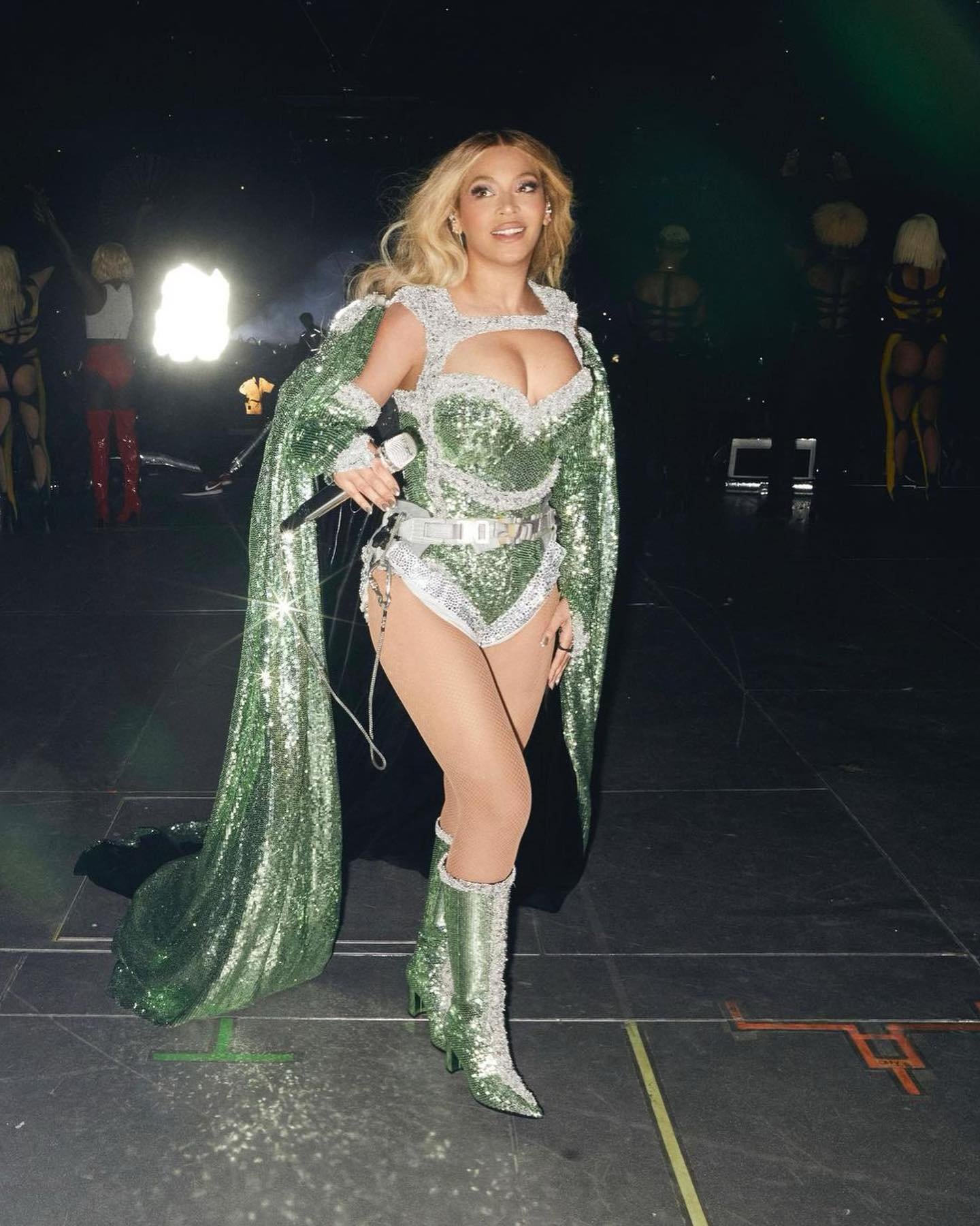 Blonde singer performs on stage in green, sparkling bodysuit with cape and knee-high boots, holding a microphone, stage lighting.
