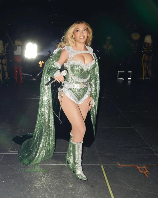 Blonde singer performs on stage in green, sparkling bodysuit with cape and knee-high boots, holding a microphone, stage lighting.