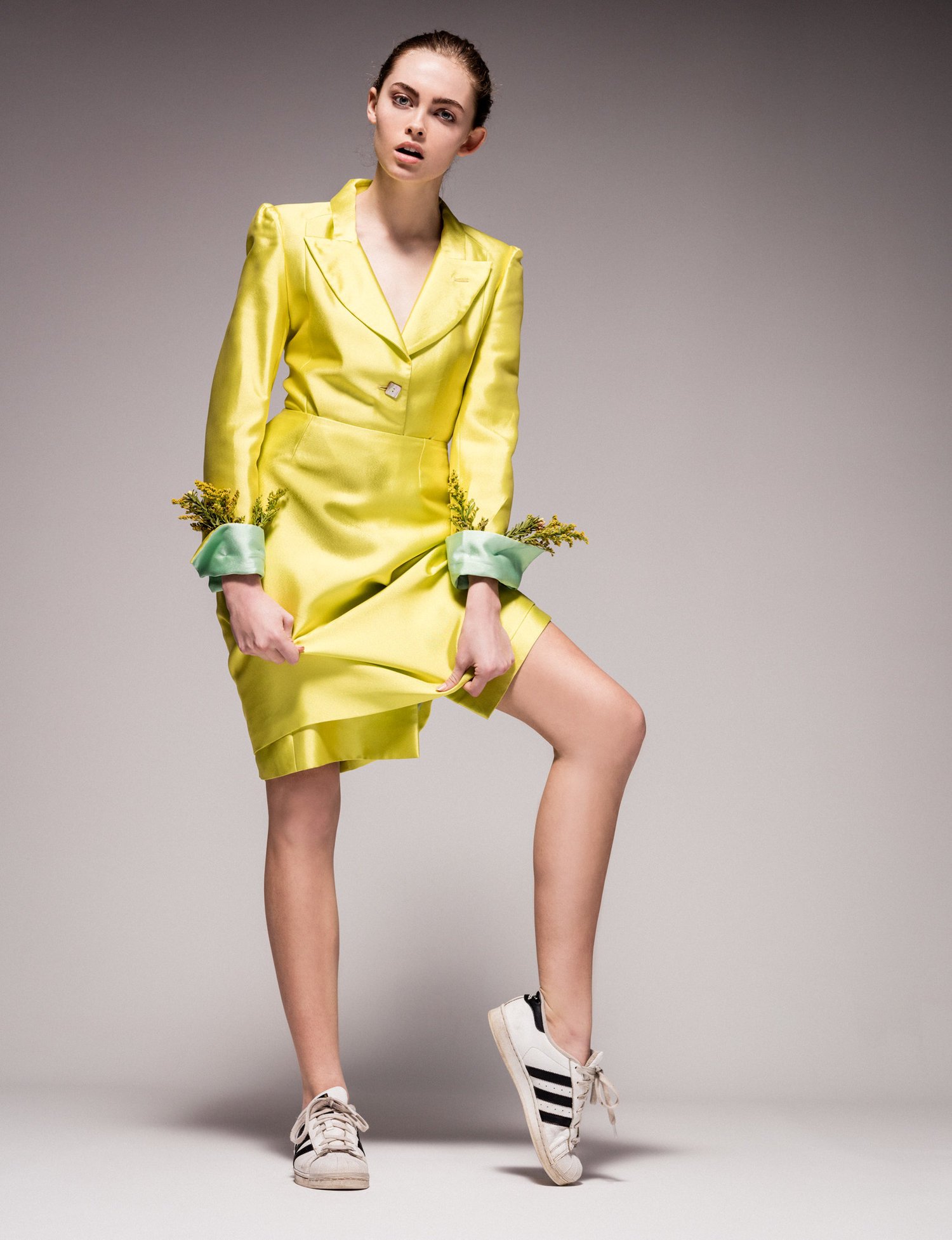 A young woman in a chartreuse skirt suit and Adidas sneakers poses in a studio against a gray background, exuding a modern and stylish look.