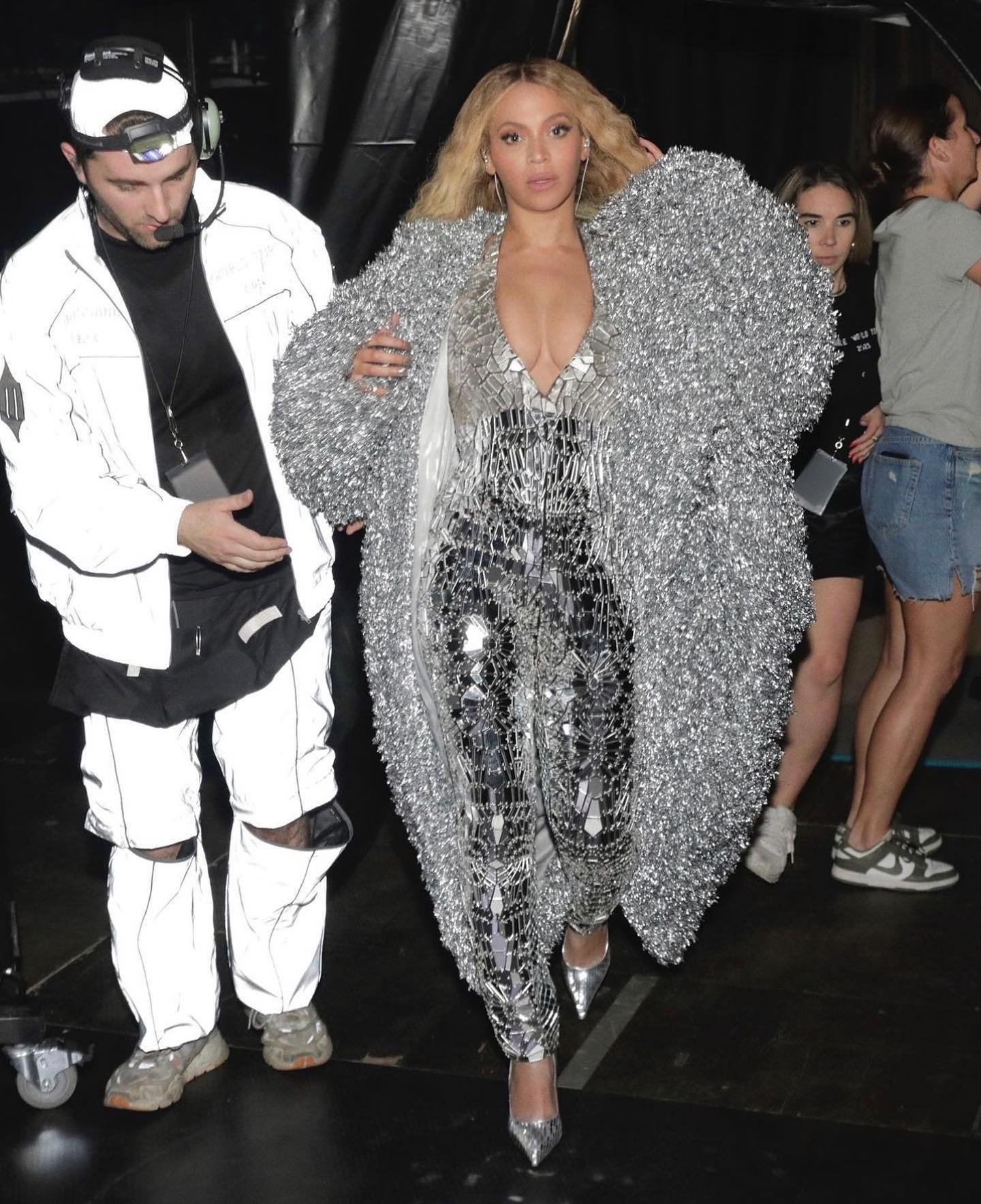 Beyonce in shimmery outfit with a silver coat, exiting with a man in reflective clothing, crowd in background, indoor, medium shot.