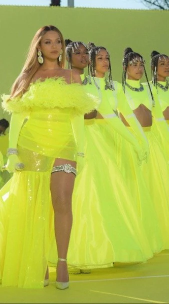Beyonce Knowles performs in yellow, alongside backup dancers in matching outfits, on an outdoor stage with green background.