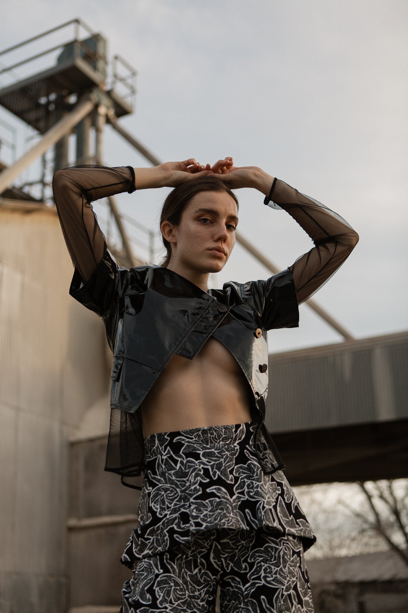 Fashionable woman poses in a black mesh top, patent leather vest, and floral print pants near an industrial building under a cloudy sky.