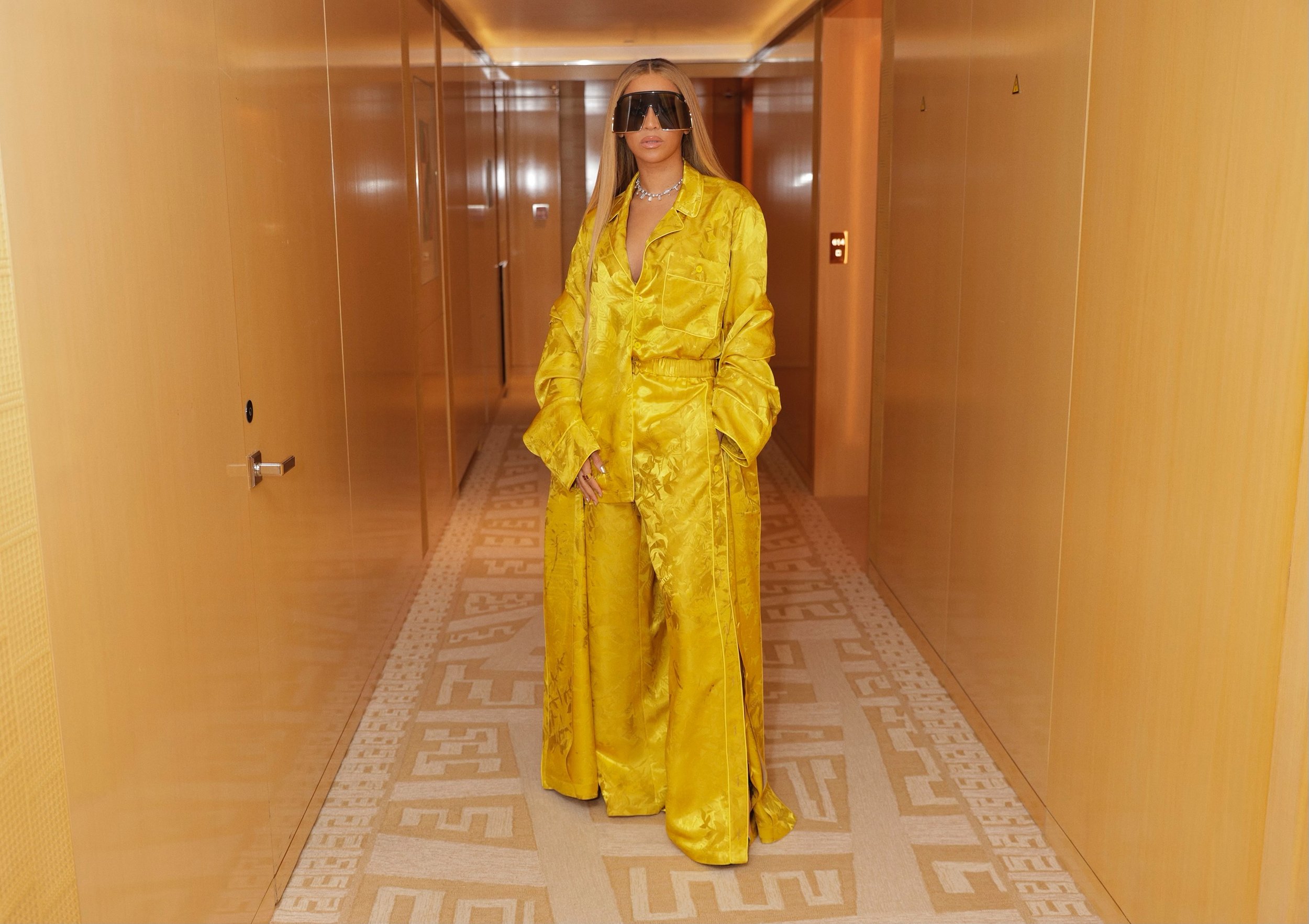 Woman in Yellow Outfit and Black Sunglasses Posing in Hallway with Ornate Carpet, Walls, and Doors.