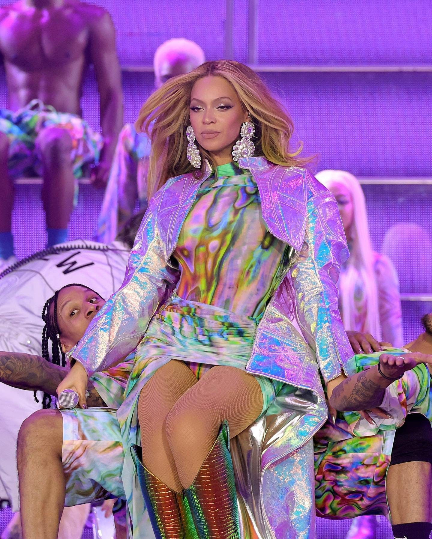 Beyonce performs on stage with dancers wearing a shimmering holographic jacket dress and sparkling jewelry in a concert setting.