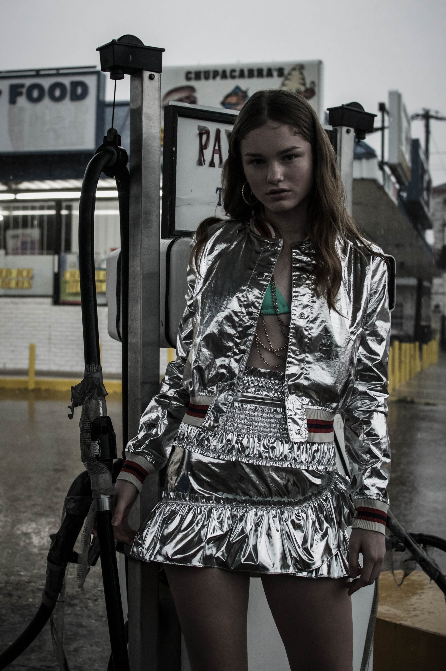 Young woman in a shiny silver skirt and jacket poses in front of gas pumps in an outdoor setting on an overcast day.
