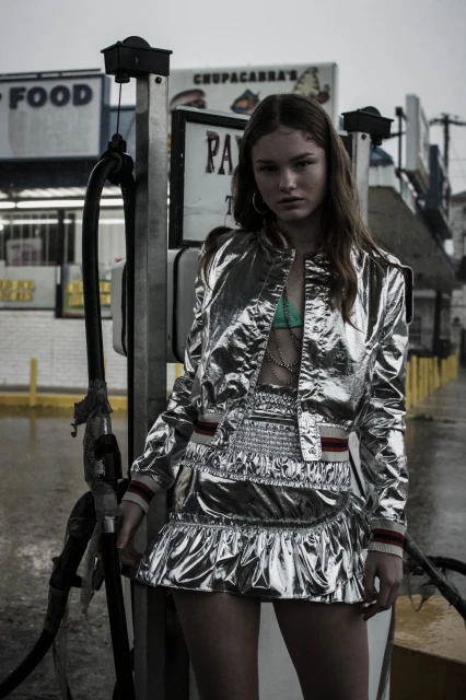Young woman in a shiny silver skirt and jacket poses in front of gas pumps in an outdoor setting on an overcast day.