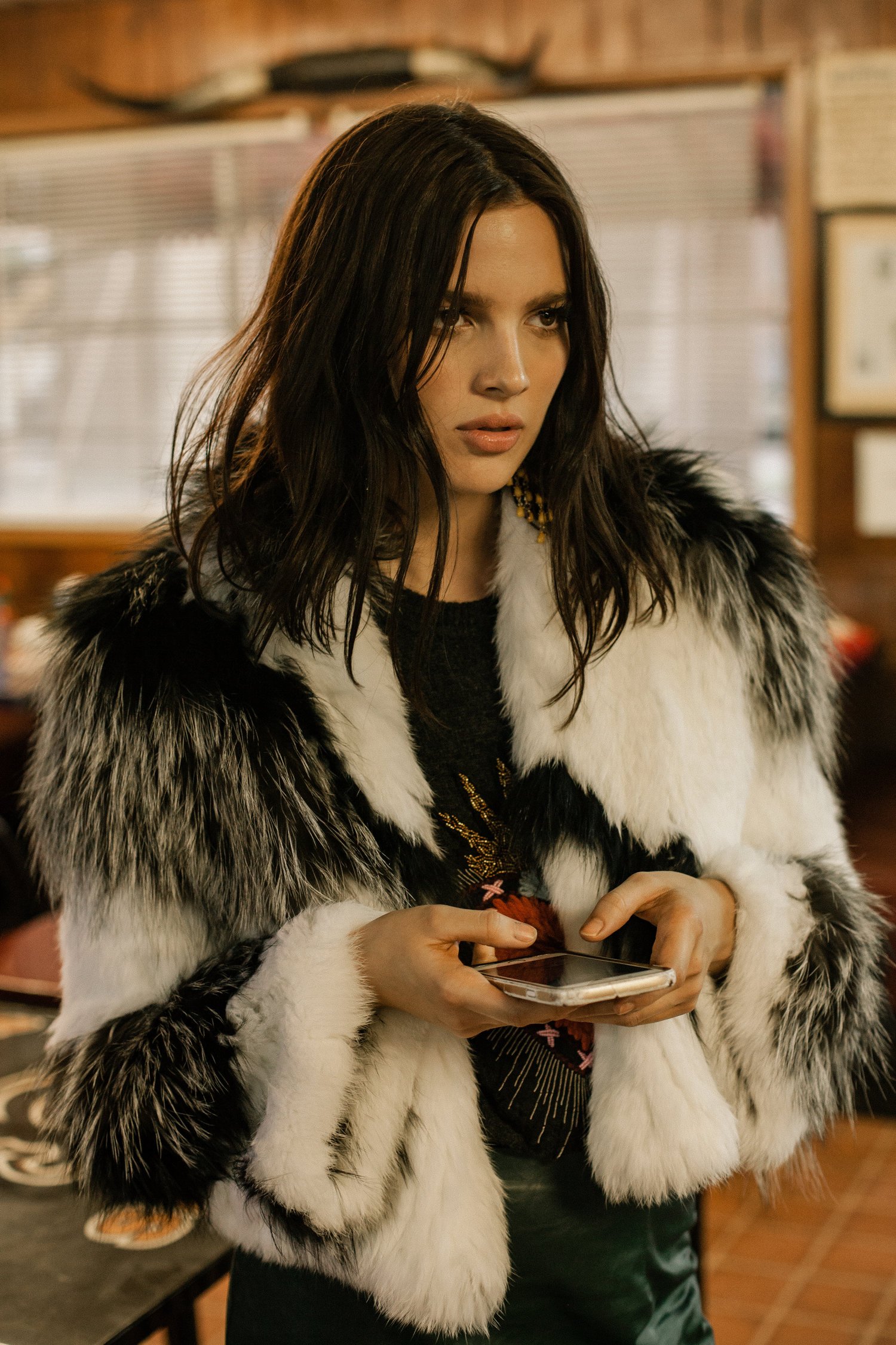 Woman in a black and white faux fur coat holding a smartphone in an interior setting.
