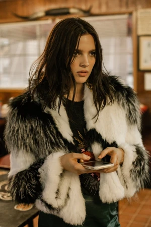 Woman in a black and white faux fur coat holding a smartphone in an interior setting.