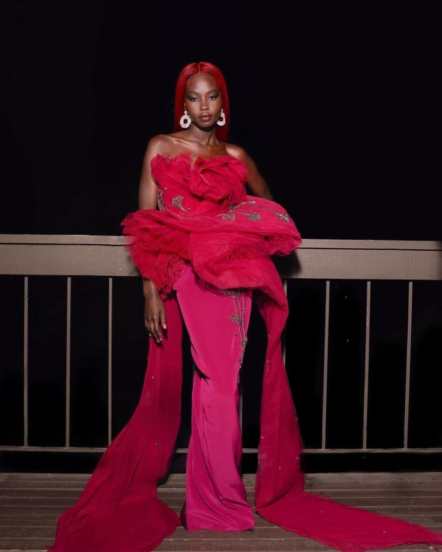 A stylish Black woman with red hair poses in a ruffled fuchsia gown with draped fabric on a wood balcony against a dark night backdrop.