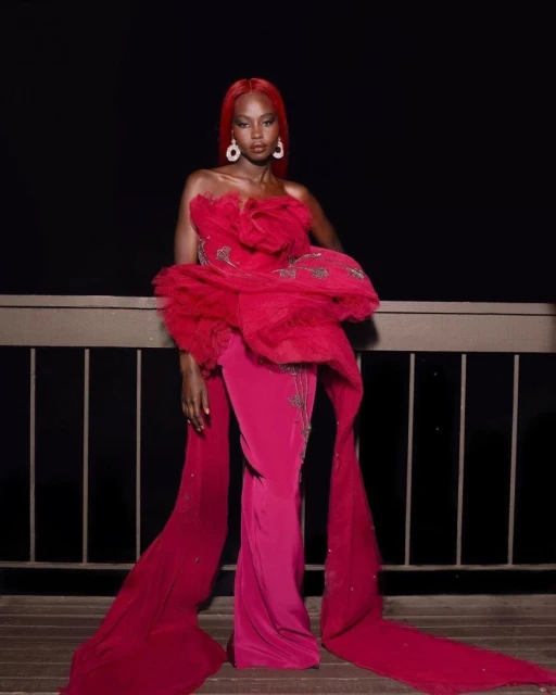 A stylish Black woman with red hair poses in a ruffled fuchsia gown with draped fabric on a wood balcony against a dark night backdrop.