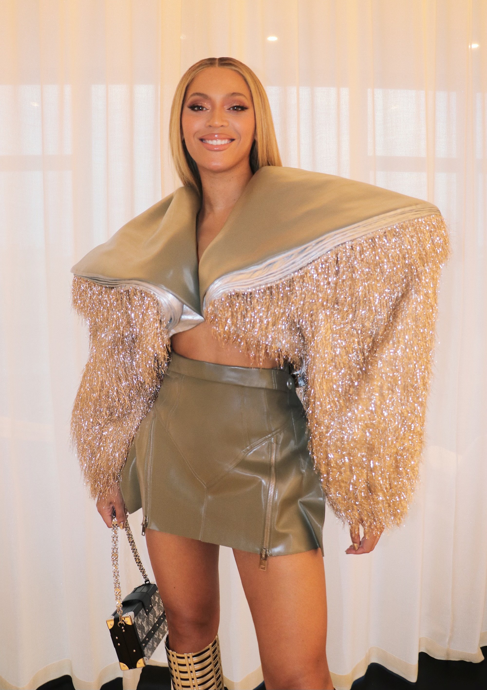 Woman in an olive green leather mini skirt and oversized jacket with gold fringe carrying a black and gold purse, smiling at the camera.