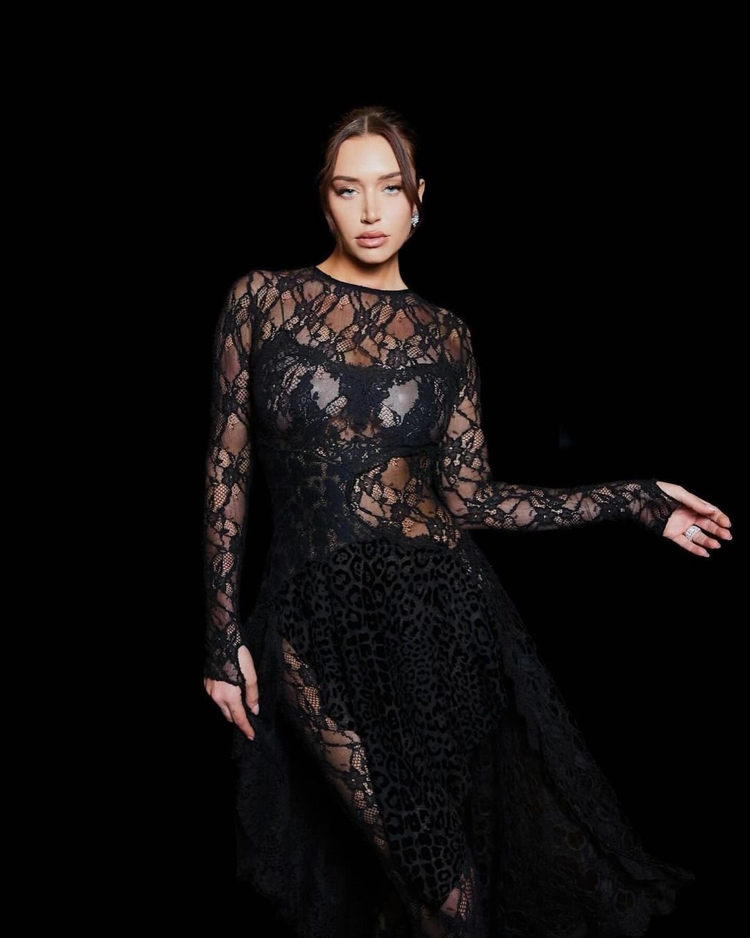 Woman in long-sleeved, semi-sheer black lace dress posing against a black backdrop in a studio. Glamour and fashion photography.