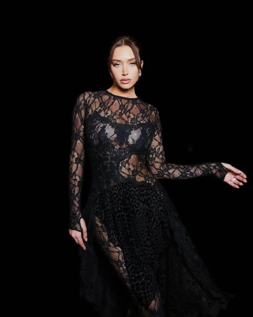 Woman in long-sleeved, semi-sheer black lace dress posing against a black backdrop in a studio. Glamour and fashion photography.