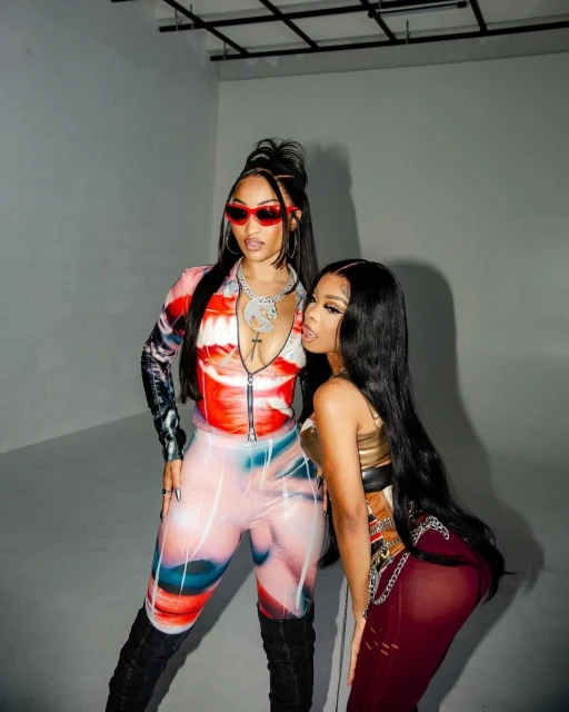 Two women posing indoors in fashionable outfits. One wears a red and white jumpsuit and the other a brown crop top and sheer skirt.
