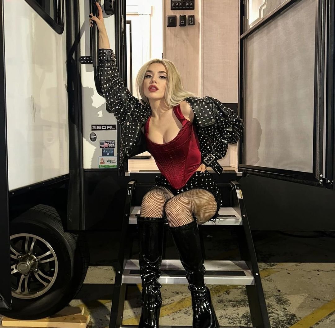 Blonde woman in a burgundy corset, polka dot jacket, and shiny black boots poses on steps of an RV with a raised hand and red lips.