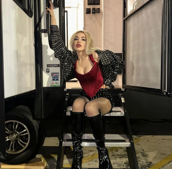 Blonde woman in a burgundy corset, polka dot jacket, and shiny black boots poses on steps of an RV with a raised hand and red lips.
